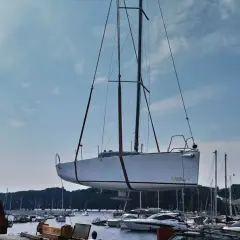 Żuraw podnosi biały jacht, którego kil jest widoczny pod kadłubem. W tle port pełen łodzi i błękitne niebo.