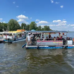 Dwa katamarany pływają po jeziorze Zegrzyńskim, przewożąc turystów. Na pierwszym planie widać łódź z rowerami i pasażerami, na drugim mniejszą łódź z żółtym daszkiem.