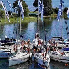 Grupa żaglówek na Mazurach, ludzie na pokładach, flagi z napisem "Na Mazury".