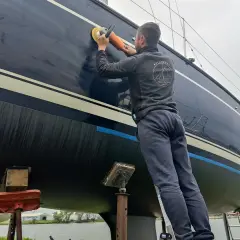 Mężczyzna poleruje lewą burtę ciemnego jachtu na stojaku.