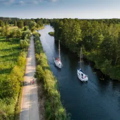 Dwa żaglowce płyną kanałem w pobliżu portu Węgorzewo. Obok ścieżka z spacerującymi ludźmi.