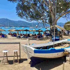 Słoneczna plaża na Hydra, Grecja, z łodzią na piasku, leżakami, parasolami i żaglówkami na morzu.
