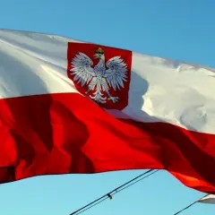 Flaga Polski z orłem w koronie powiewa na wietrze. Biało-czerwony znak bandery dumnie prezentuje się na tle błękitnego nieba.
