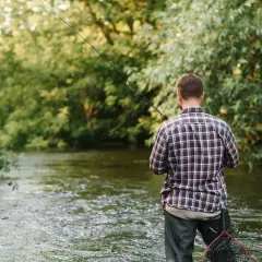 Mężczyzna w kratę łowi ryby w rzece, trzymając wędkę. W tle bujna zieleń drzew. To jego przykosa na rzece.