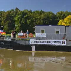 Barka z kontenerami płynie po wodach śródlądowych. Na pokładzie ludzie, flagi i baner "ODZIERAMY ODRĘ Z MITÓW".