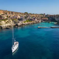 Malta: jakie morze! Biały żaglowiec kołysze się na turkusowych wodach zatoki, w tle kolorowe domki i klify.