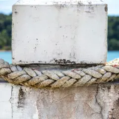 Gruba, pleciona lina, idealna do mocowania lin na jachcie, opasana wokół betonowego słupka. W tle błękitna woda i zieleń.