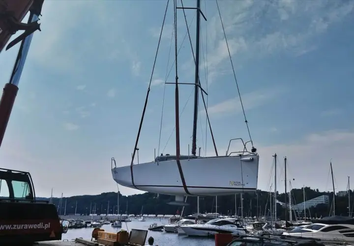 Żuraw podnosi biały jacht, którego kil jest widoczny pod kadłubem. W tle port pełen łodzi i błękitne niebo.