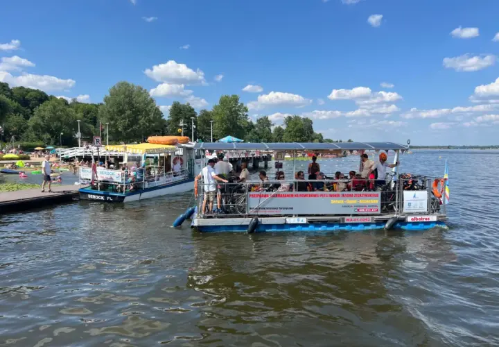Dwa katamarany pływają po jeziorze Zegrzyńskim, przewożąc turystów. Na pierwszym planie widać łódź z rowerami i pasażerami, na drugim mniejszą łódź z żółtym daszkiem.