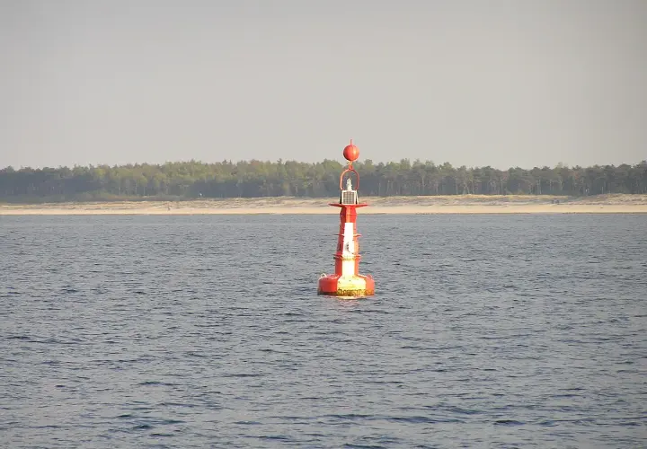 Czerwono-biała boja nawigacyjna, znak bezpiecznej wody, na spokojnym morzu. W tle linia lasu i plaży.