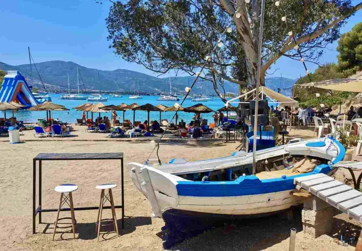 Słoneczna plaża na Hydra, Grecja, z łodzią na piasku, leżakami, parasolami i żaglówkami na morzu.