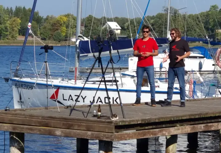 Dwóch mężczyzn na pomoście nagrywa film na tle żaglówki "Lazy Jack". Na pomoście leży kot.