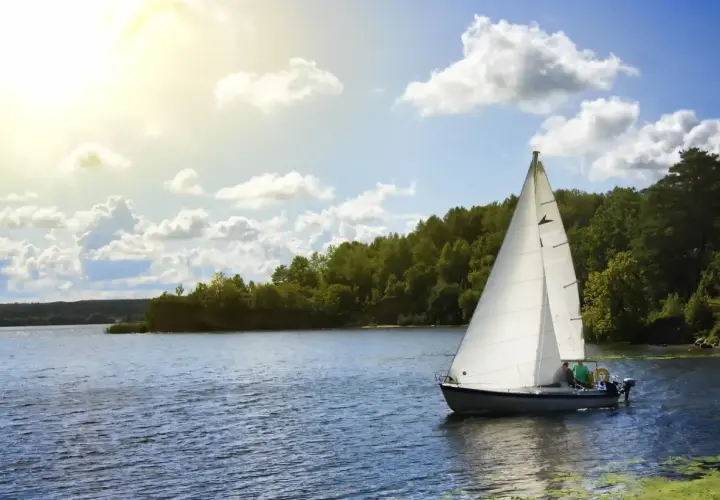 Ahoj Mazury! Biały żagiel łodzi odbija się w spokojnej tafli jeziora, otoczonej zielonymi lasami. Słońce przebija się przez chmury.