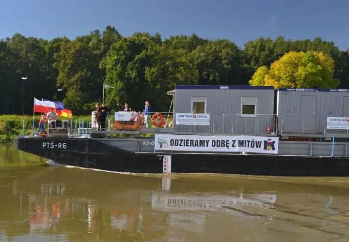 Barka z kontenerami płynie po wodach śródlądowych. Na pokładzie ludzie, flagi i baner "ODZIERAMY ODRĘ Z MITÓW".