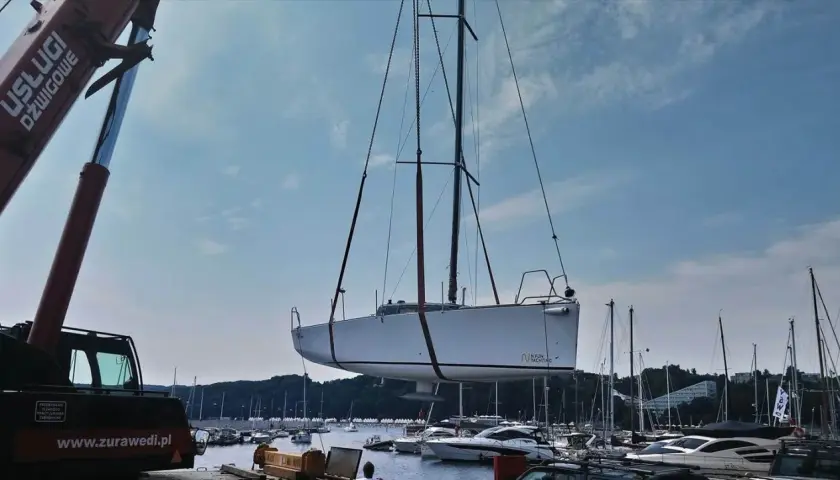 Żuraw podnosi biały jacht, którego kil jest widoczny pod kadłubem. W tle port pełen łodzi i błękitne niebo.