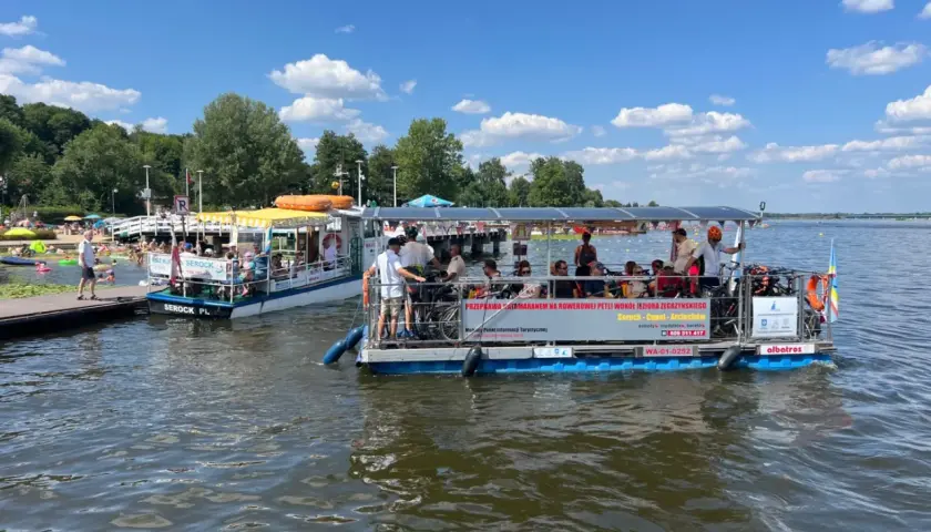 Dwa katamarany pływają po jeziorze Zegrzyńskim, przewożąc turystów. Na pierwszym planie widać łódź z rowerami i pasażerami, na drugim mniejszą łódź z żółtym daszkiem.