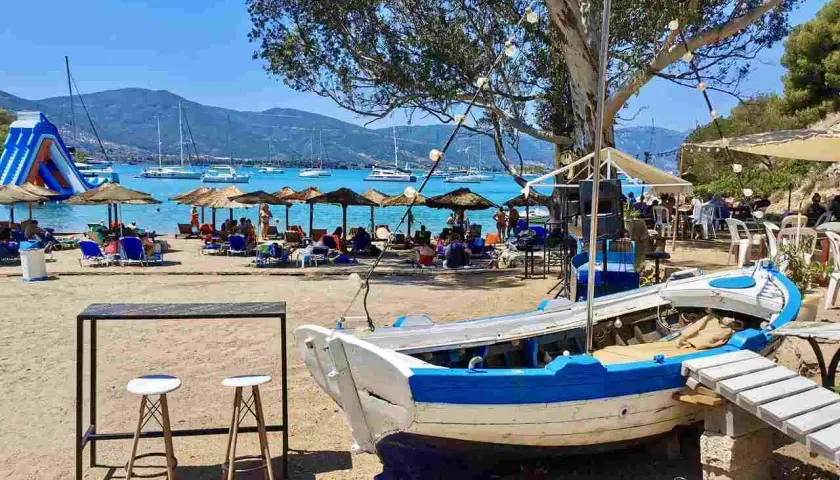 Słoneczna plaża na Hydra, Grecja, z łodzią na piasku, leżakami, parasolami i żaglówkami na morzu.