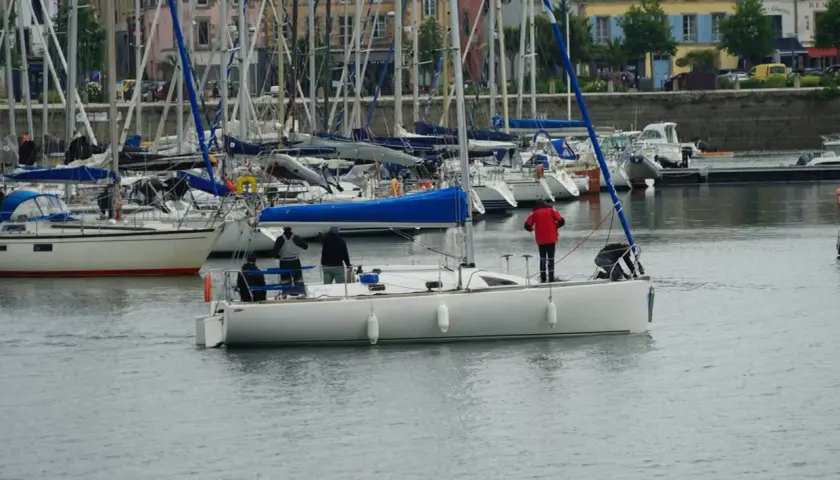 Załoga na jachcie przygotowuje się do manewru, jeden z marynarzy stoi przy sterze, inny pochyla się nad pokładem. W tle port z wieloma żaglówkami i budynkami. Szpring rufowy jest niewidoczny.