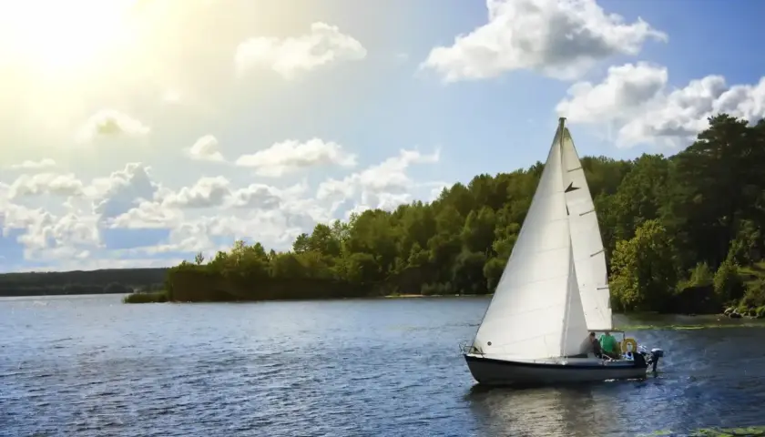 Ahoj Mazury! Biały żagiel łodzi odbija się w spokojnej tafli jeziora, otoczonej zielonymi lasami. Słońce przebija się przez chmury.