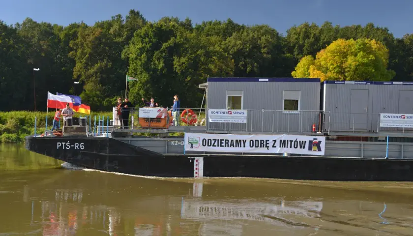 Barka z kontenerami płynie po wodach śródlądowych. Na pokładzie ludzie, flagi i baner "ODZIERAMY ODRĘ Z MITÓW".