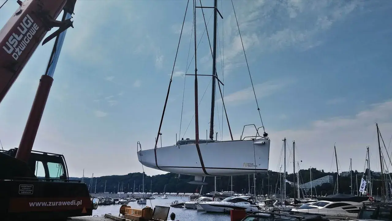 Żuraw podnosi biały jacht, którego kil jest widoczny pod kadłubem. W tle port pełen łodzi i błękitne niebo.