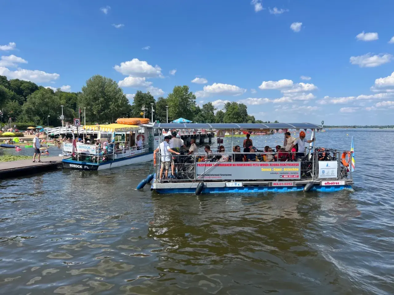 Dwa katamarany pływają po jeziorze Zegrzyńskim, przewożąc turystów. Na pierwszym planie widać łódź z rowerami i pasażerami, na drugim mniejszą łódź z żółtym daszkiem.