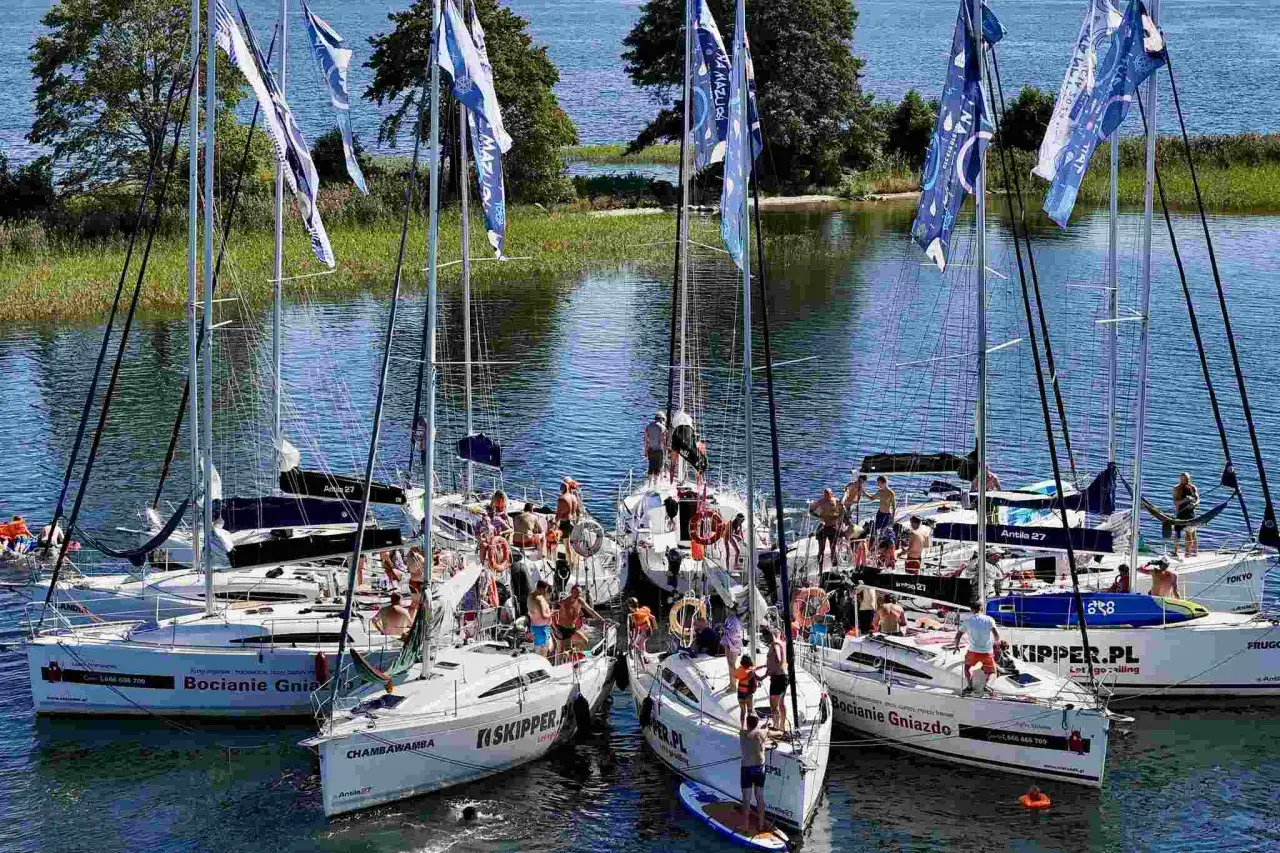 Grupa żaglówek na Mazurach, ludzie na pokładach, flagi z napisem "Na Mazury".