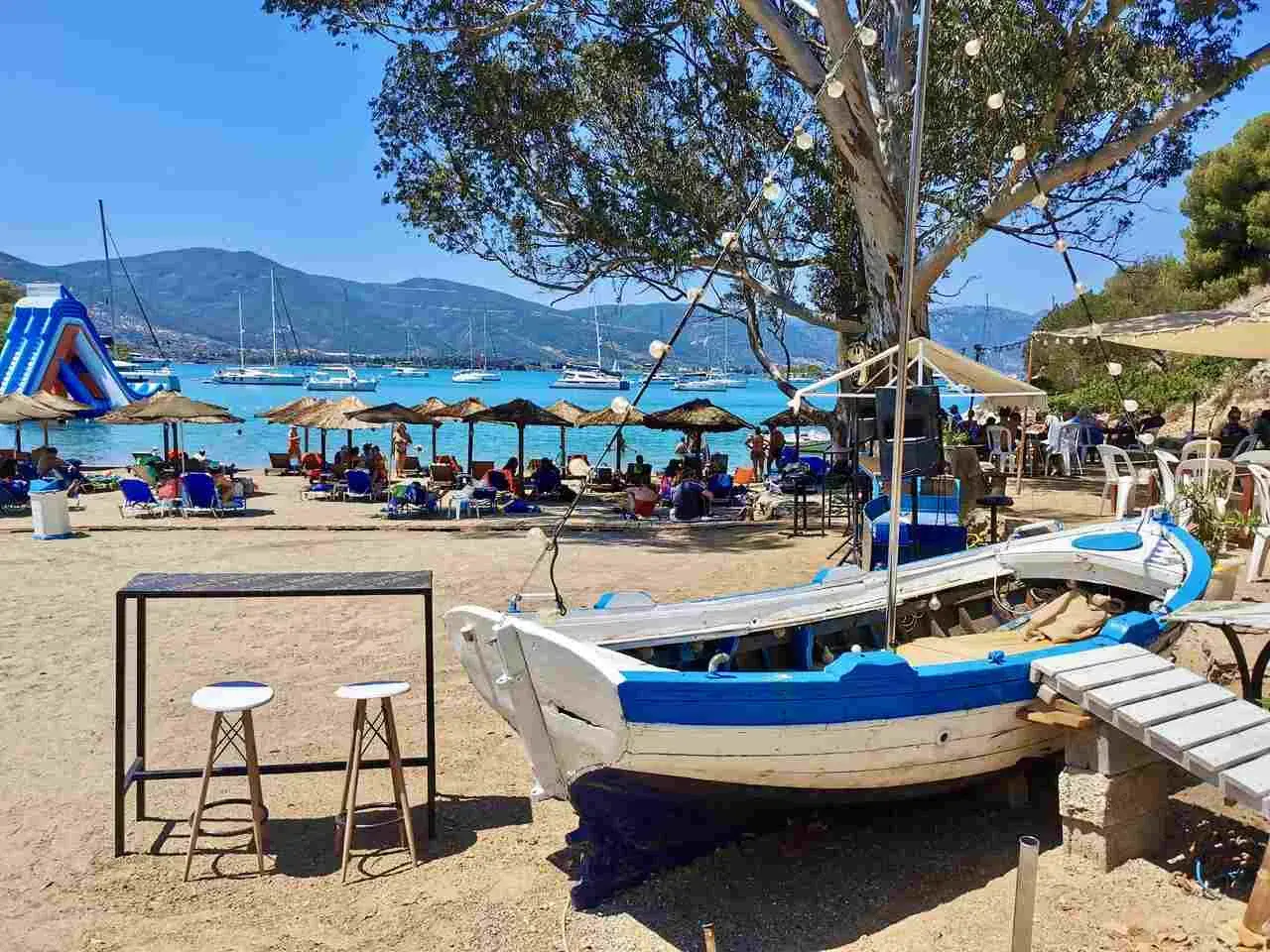 Słoneczna plaża na Hydra, Grecja, z łodzią na piasku, leżakami, parasolami i żaglówkami na morzu.