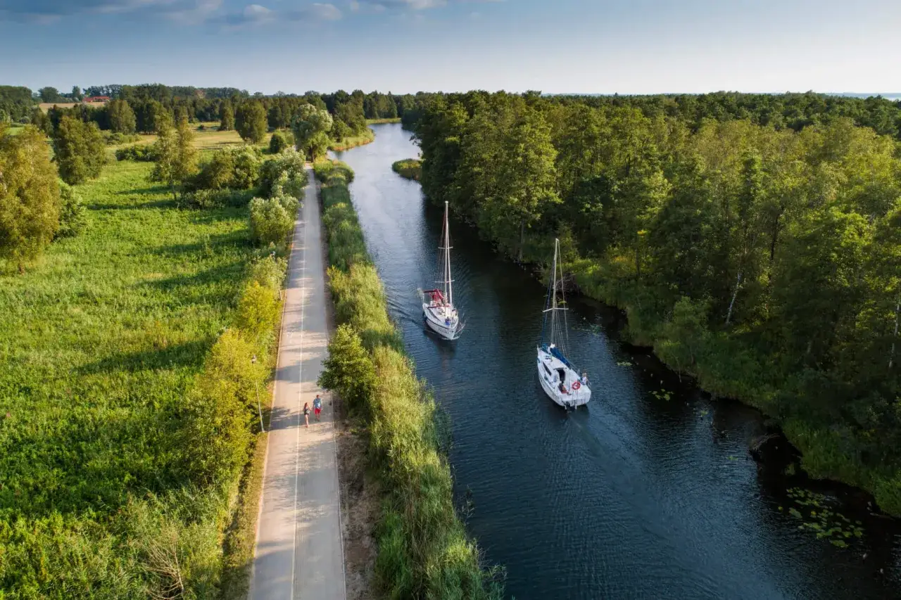 Dwa żaglowce płyną kanałem otoczonym zielenią. Obok ścieżka, którą spaceruje para. Piękny brzeg Mazury.